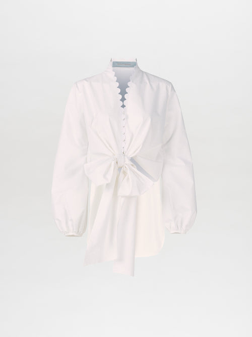 The Honey Blouse White Eyelet showcases a long-sleeve design in white, complete with scalloped edges, a button-up front, and a distinctive neckline highlighted by a large front bow. Made from soft cotton, this blouse seamlessly combines elegance with comfort.