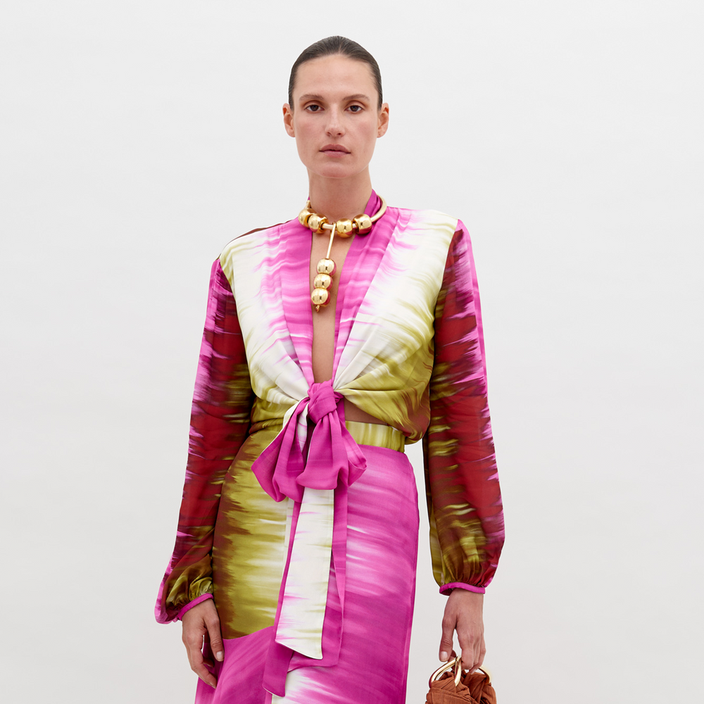 A woman with tied-back hair wears a Honey Blouse Violeta Lime Tie Dye with a front knot and matching skirt, accessorized with a gold bead necklace and holding a brown handbag against a plain background. This pre-order item will ship by November 15th, 2024.
