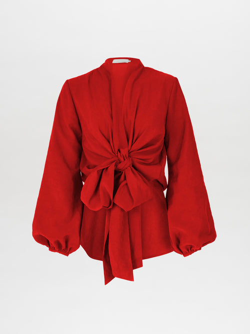 The Honey Blouse Rouge is a vibrant rouge fabric blouse featuring a red long-sleeved design with a large front knot, bishop sleeves with elastic cuffs, and a plunging neckline.