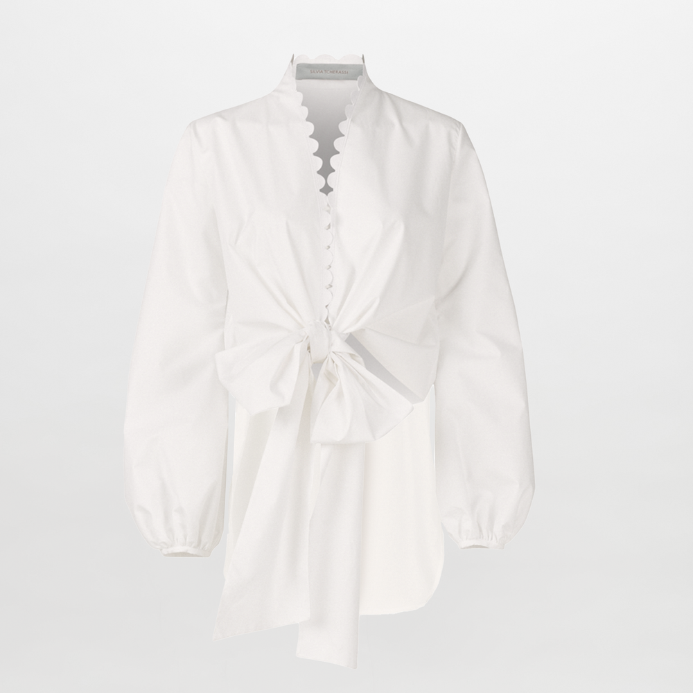 The Honey Blouse White Eyelet features white cotton with organza sleeves, scalloped edges, a distinctive neckline, front bow detail, and an asymmetrical hem, shown against a plain white background.