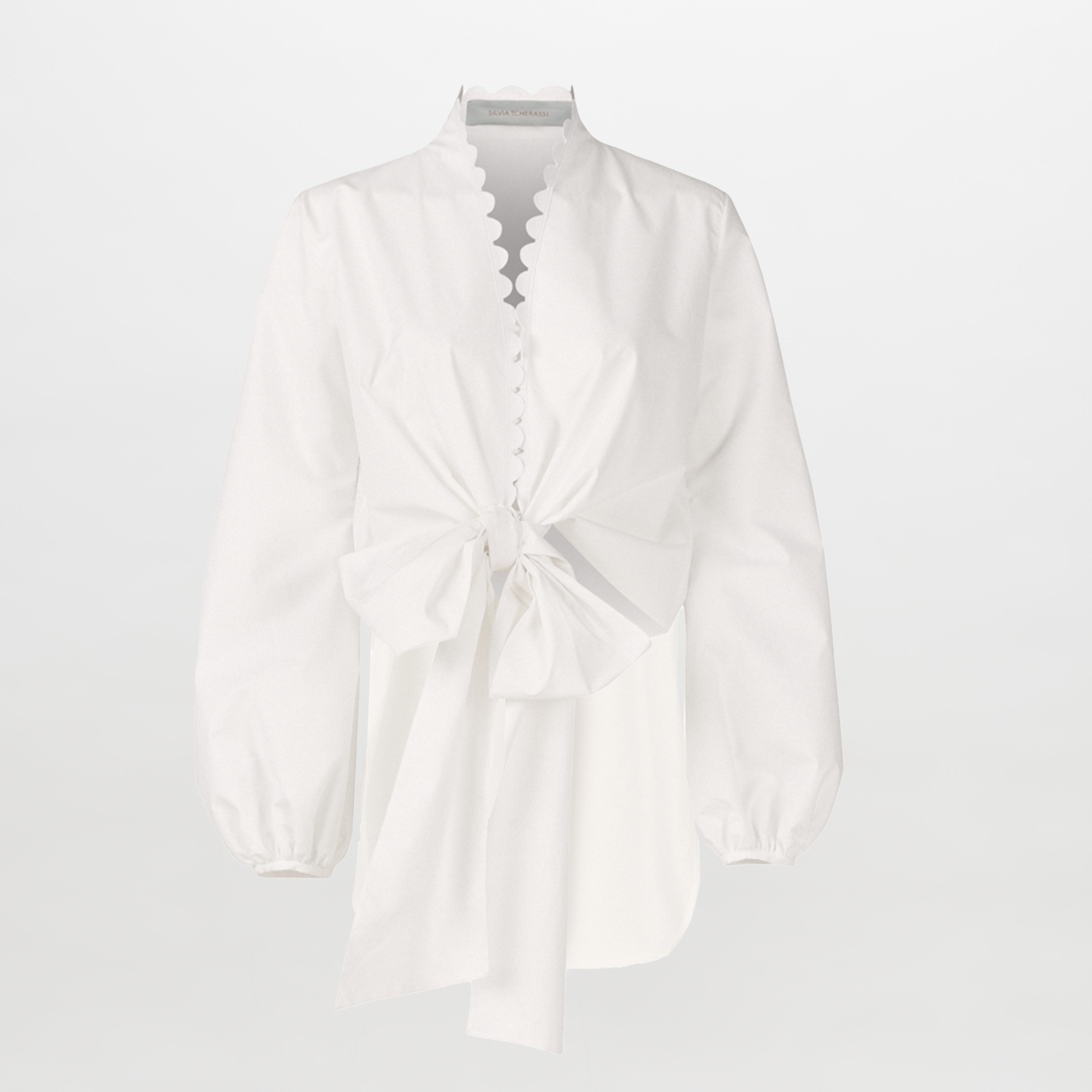 The Honey Blouse White Eyelet features white cotton with organza sleeves, scalloped edges, a distinctive neckline, front bow detail, and an asymmetrical hem, shown against a plain white background.