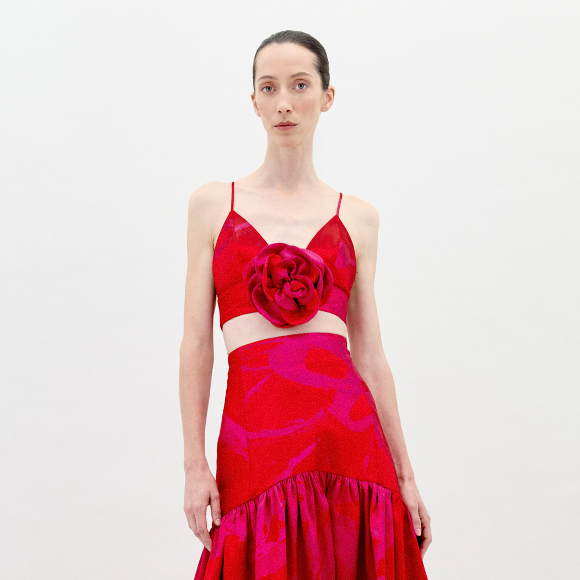 A person stands against a plain background wearing the Ibbie Top Rouge Fuchsia Flower, a red taffeta crop top with thin straps and a large floral appliqué on the front, paired with a skirt featuring gathered fabric at the hem.
