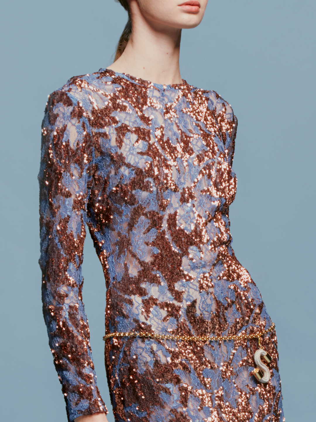 Person in the Ida Dress Navy Ochre Sequin, a long-sleeved, floor-length gown featuring an intricate blue and brown pattern, stands against a plain blue background, ready to dazzle at the gala on September 2nd.