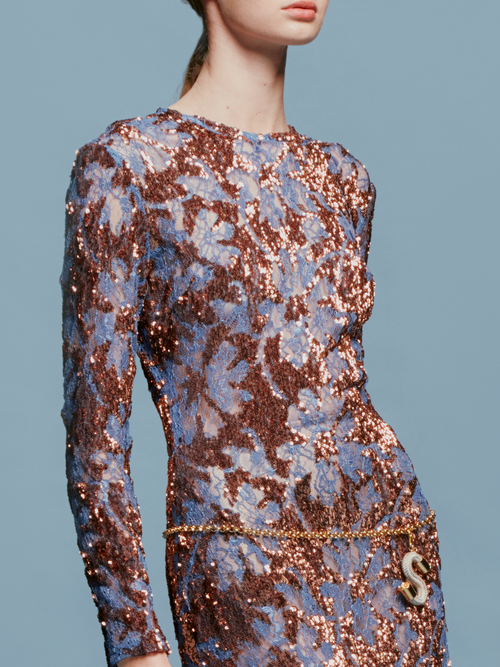 Person in the Ida Dress Navy Ochre Sequin, a long-sleeved, floor-length gown featuring an intricate blue and brown pattern, stands against a plain blue background, ready to dazzle at the gala on September 2nd.