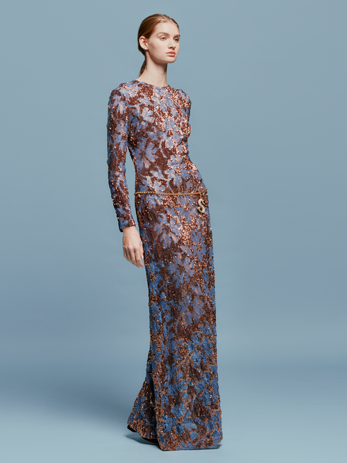 Person in the Ida Dress Navy Ochre Sequin, a long-sleeved, floor-length gown featuring an intricate blue and brown pattern, stands against a plain blue background, ready to dazzle at the gala on September 2nd.