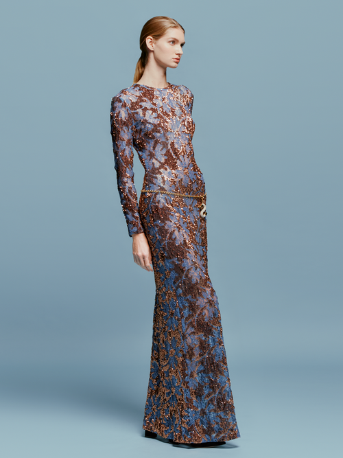 Person in the Ida Dress Navy Ochre Sequin, a long-sleeved, floor-length gown featuring an intricate blue and brown pattern, stands against a plain blue background, ready to dazzle at the gala on September 2nd.