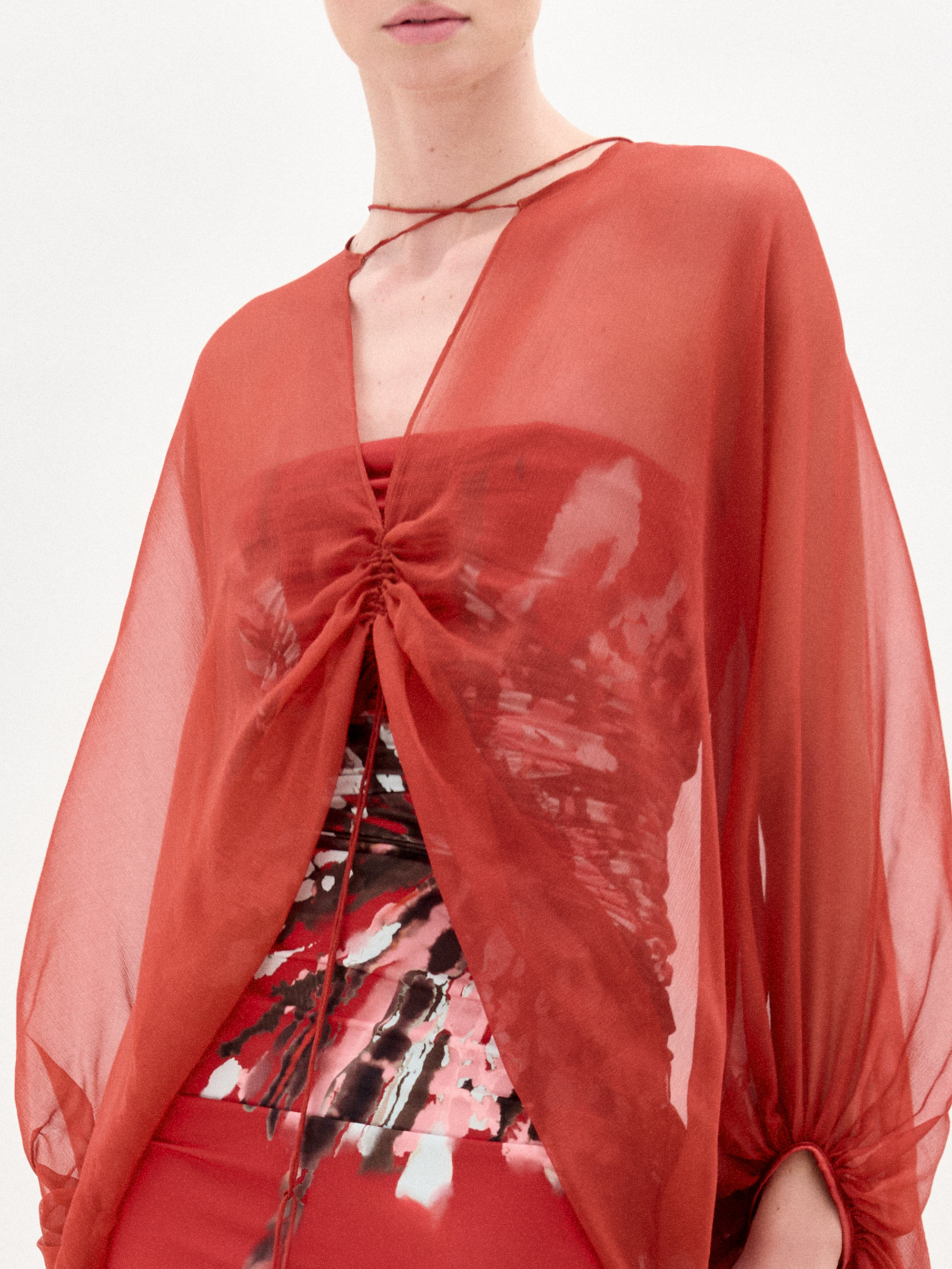A woman stands against a plain background wearing the Idra Blouse, a sheer, long-sleeved top with a deep V neckline, layered over a patterned shirt and paired with a matching red skirt.