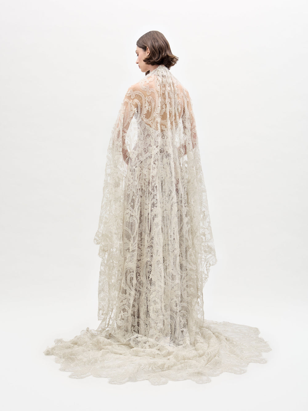 A person clad in an Olinta Wedding Veil holds a floral bouquet. The intricate lace gown boasts a high neckline, a cape, and is set against a plain white background.