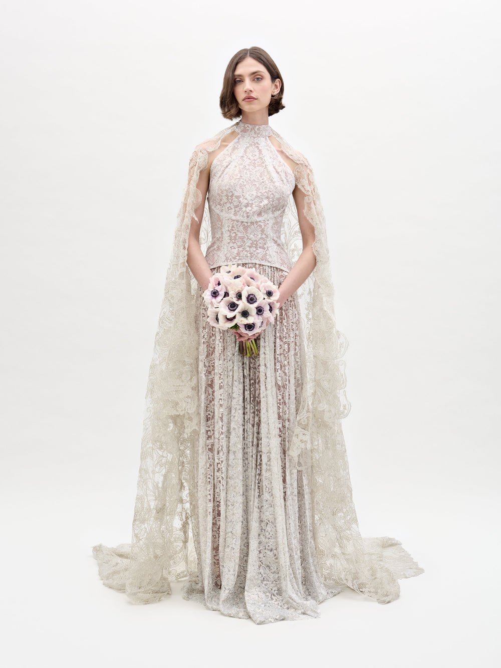 Wearing the Ilana Dress, a woman in a long lace bridal gown holds a bouquet of white and purple flowers against a plain background.
