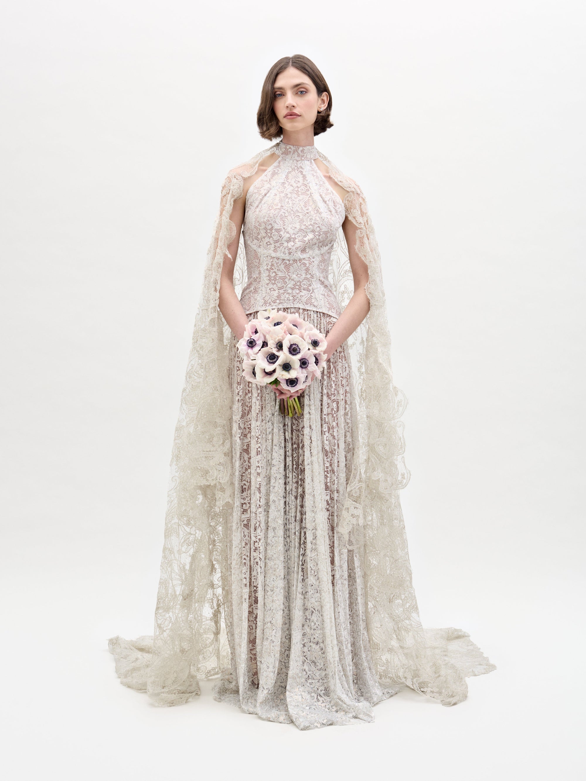 Wearing the Ilana Dress, a woman in a long lace bridal gown holds a bouquet of white and purple flowers against a plain background.