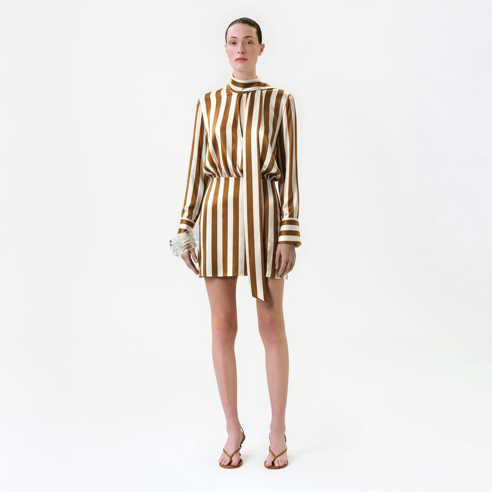 A person stands against a white background wearing the Imelda Dress, a brown and white vertically striped silk mini dress, paired with flip-flop sandals and holding a small textured accessory crafted from fine Italian fabric.