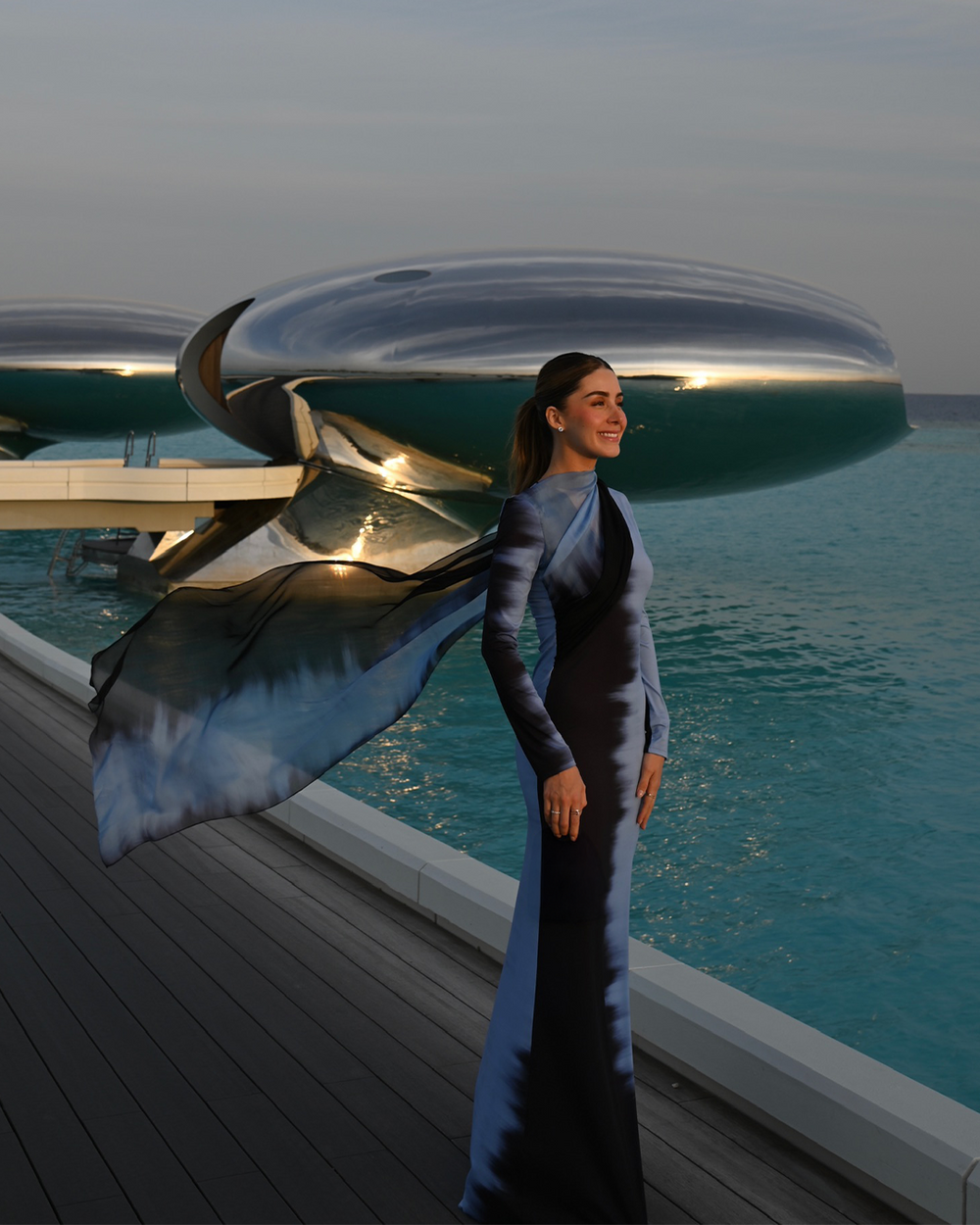 Woman in a blue dress standing on a deck with futuristic architecture and ocean view.