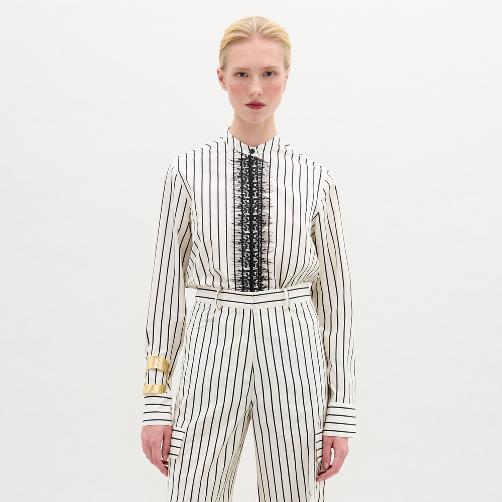 A person stands against a plain white background wearing the Isabell Blouse, a white long-sleeve top with vertical black stripes and intricate black lace embroidery.