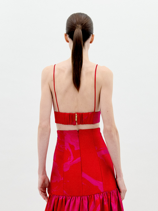 A person stands against a plain background wearing the Ibbie Top Rouge Fuchsia Flower, a red taffeta crop top with thin straps and a large floral appliqué on the front, paired with a skirt featuring gathered fabric at the hem.