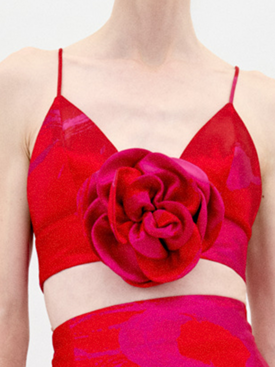 A person stands against a plain background wearing the Ibbie Top Rouge Fuchsia Flower, a red taffeta crop top with thin straps and a large floral appliqué on the front, paired with a skirt featuring gathered fabric at the hem.