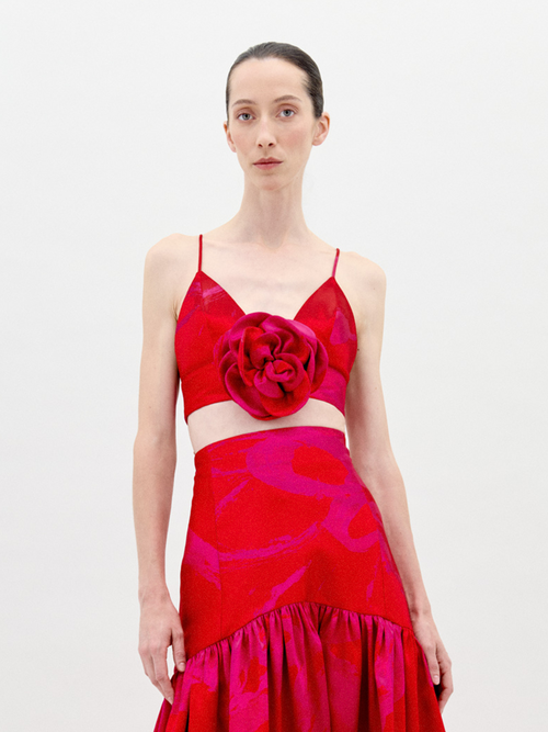 A person stands against a plain background wearing the Ibbie Top Rouge Fuchsia Flower, a red taffeta crop top with thin straps and a large floral appliqué on the front, paired with a skirt featuring gathered fabric at the hem.