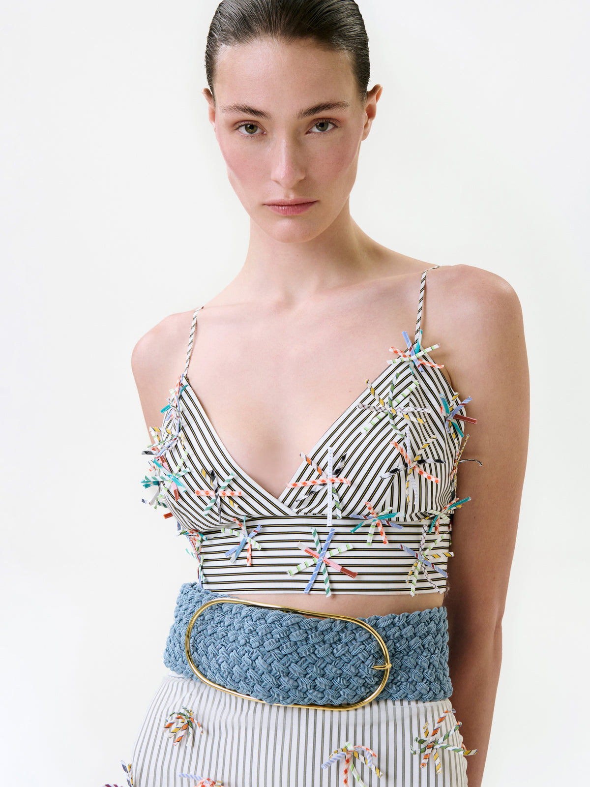 Ibbie Top, a cropped V-neck with thin straps, vertical stripes, and colorful fringe accents, paired with a wide blue woven belt featuring a gold buckle.