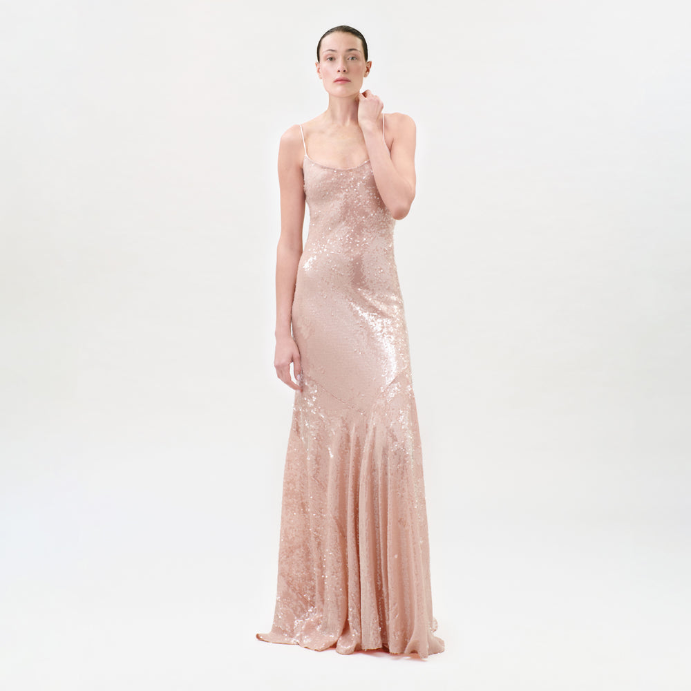 A woman stands against a plain white background wearing the Ibona Dress, a sleeveless, floor-length maxi gown in light pink sequins, crafted from luxurious Italian fabric. Her hair is pulled back and one hand gently touches her neck.