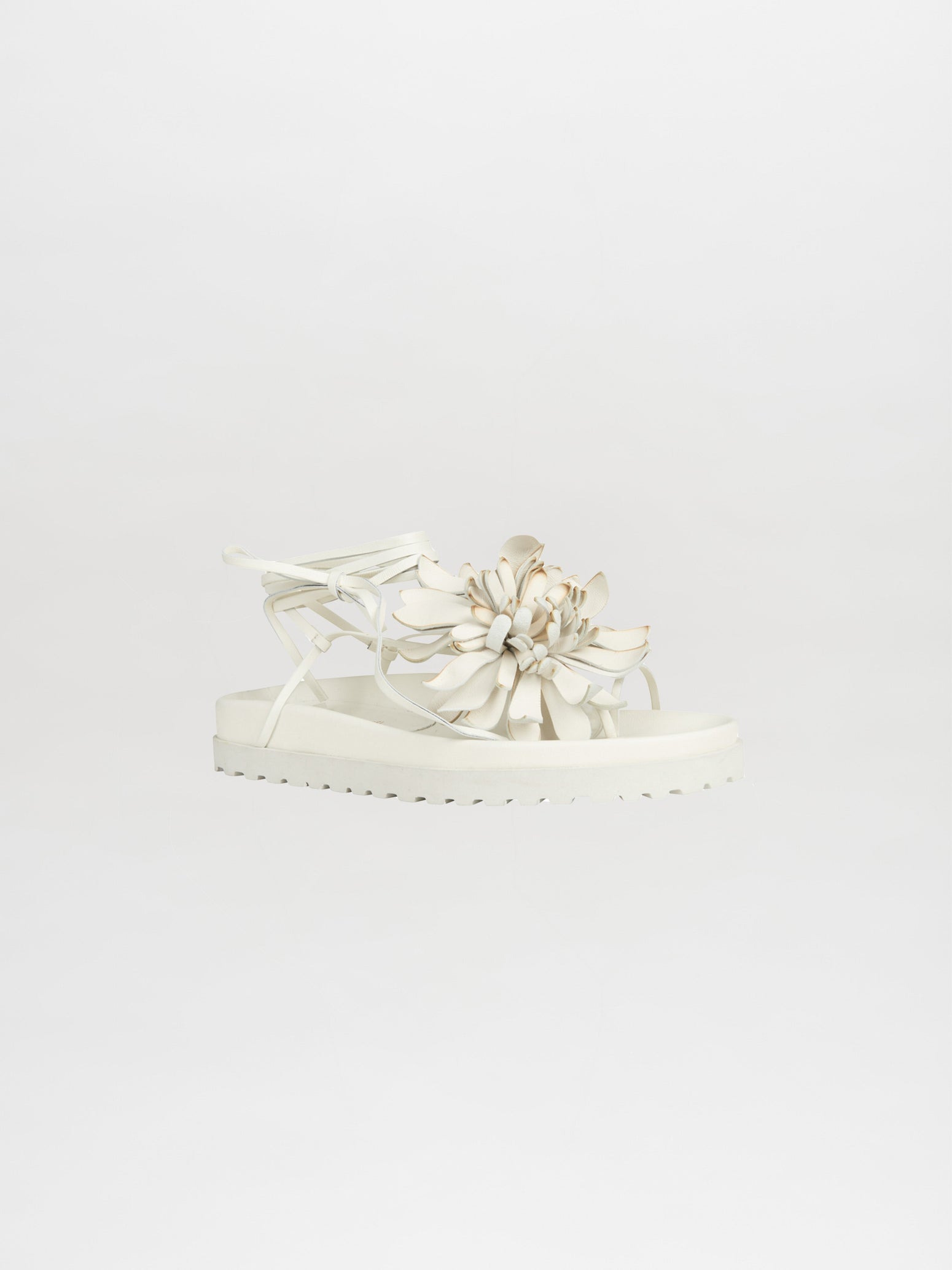 Idania Sandal Beige featuring a large floral decoration on top and intricate ankle straps, available for shipping within two weeks. Photographed against a plain background.