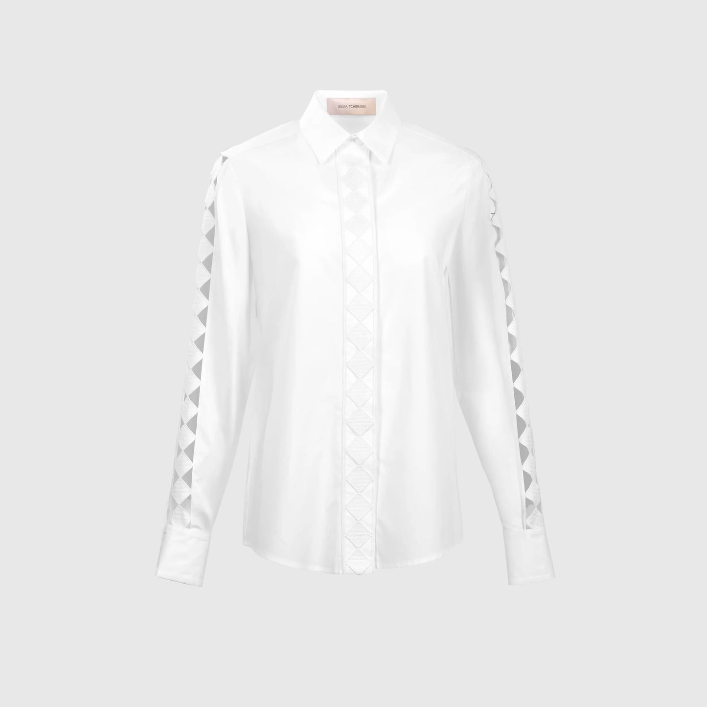 The Idris Blouse is a white long-sleeve top with a pointed collar, made from quality Portuguese fabric. It showcases a geometric triangular pattern on the sleeves and front placket, set against a neutral gray background.