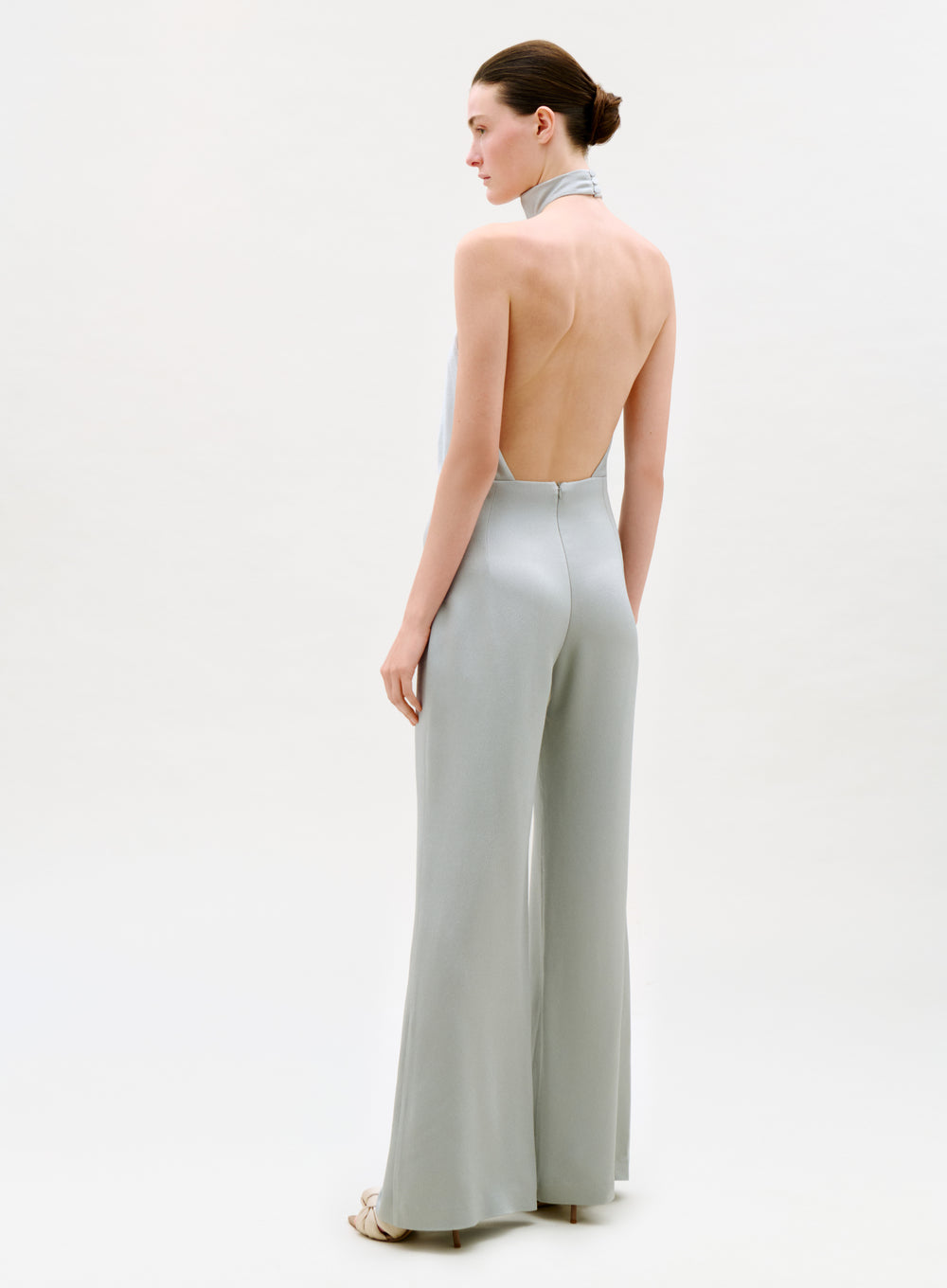 A woman stands against a plain background wearing the Imperio Jumpsuit—a light gray, backless halter style with wide-leg pants and high heels. She is turned to the side, highlighting its open-back design.