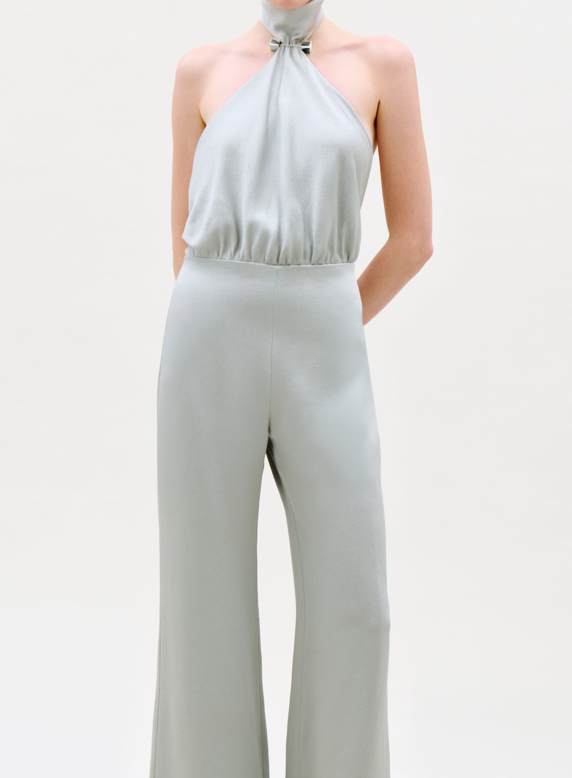 Someone stands against a plain light background wearing the Imperio Jumpsuit, a sleeveless, light gray halter-neck style with wide-leg pants, gathered neckline, and a fitted waist.