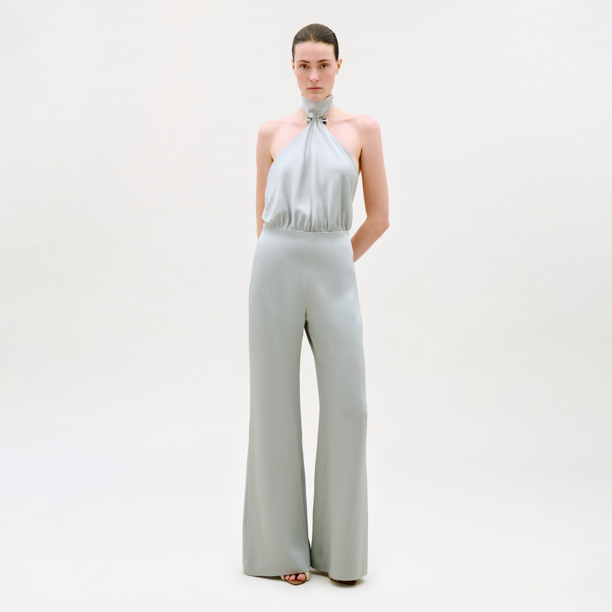 A woman stands against a plain light background wearing the Imperio Jumpsuit—a light gray, sleeveless halter-neck design with wide-leg pants. Her hair is pulled back and her hands are behind her back.
