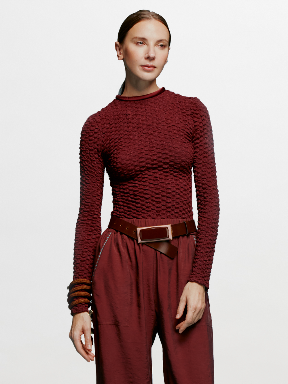 A person with long hair is wearing the Jari Top Burgundy, matching pants, and a wide belt, standing against a blue background like they're ready to set sail on a ship.