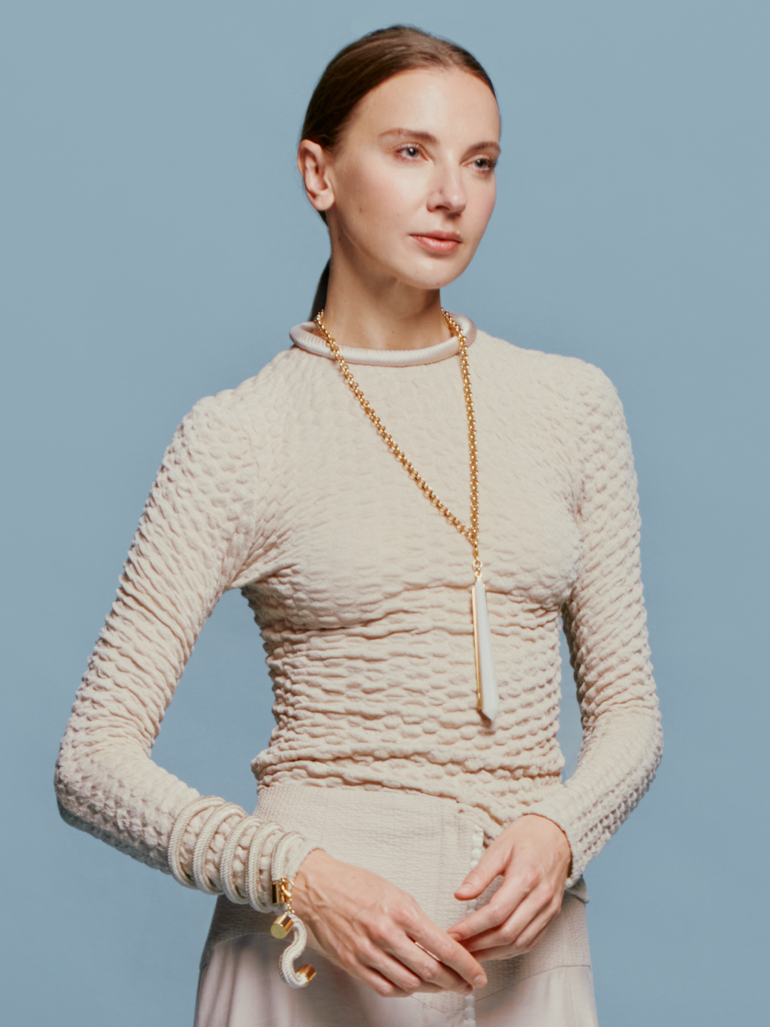 A woman with tied-back hair wears the Jari Top Cream, a long gold necklace, and several bracelets, standing against a light blue background reminiscent of the sky on August 19th.