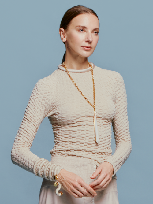 A woman with tied-back hair wears the Jari Top Cream, a long gold necklace, and several bracelets, standing against a light blue background reminiscent of the sky on August 19th.