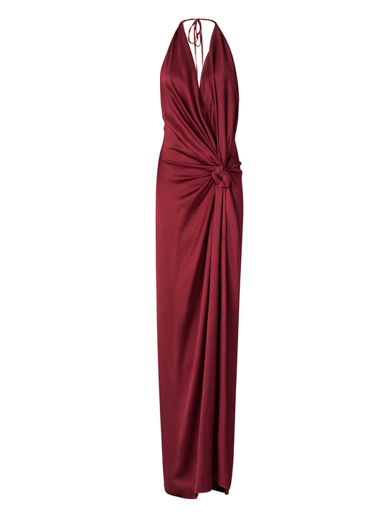 A woman wearing the Jazmin Dress Burgundy poses against a blue background. This long, sleeveless dress features a halter neckline and draping detail, perfect for making an entrance on a glamorous August 19th evening.