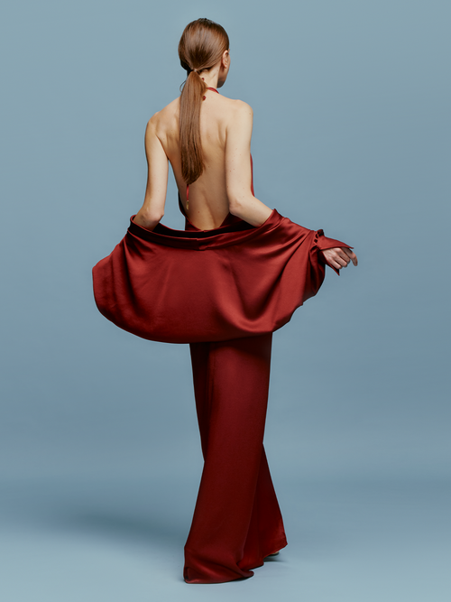 A woman wearing the Jazmin Dress Burgundy poses against a blue background. This long, sleeveless dress features a halter neckline and draping detail, perfect for making an entrance on a glamorous August 19th evening.