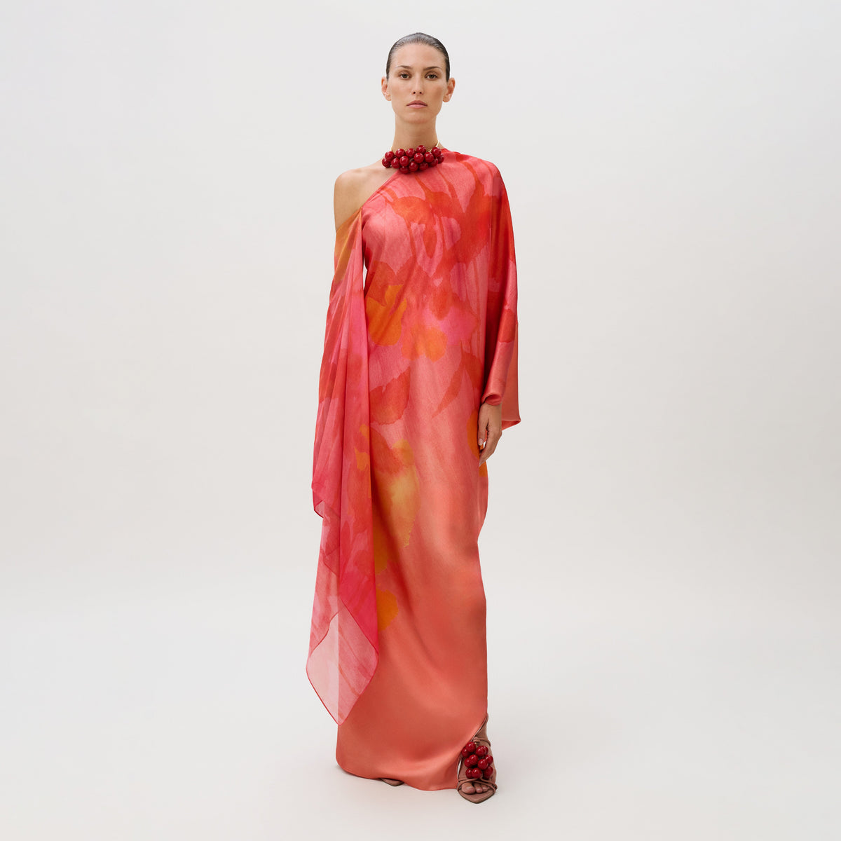 A model wears the Jade Dress, a floor-length, one-shoulder silk chiffon gown in coral and pink with a draped side panel, styled with a red beaded collar and matching beaded sandals.