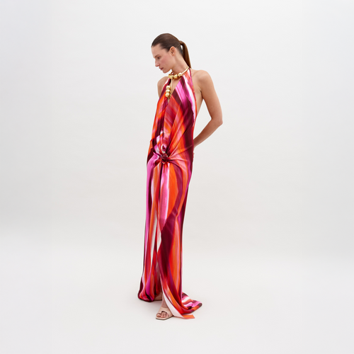 A woman stands against a plain background, wearing a colorful, flowing halter-neck Jazmin Dress Magenta Abstract Waves with a prominent knot detail at the waist. Available for pre-order now, the dress will ship by November 15th, 2024. She has her hands behind her back and is looking down.