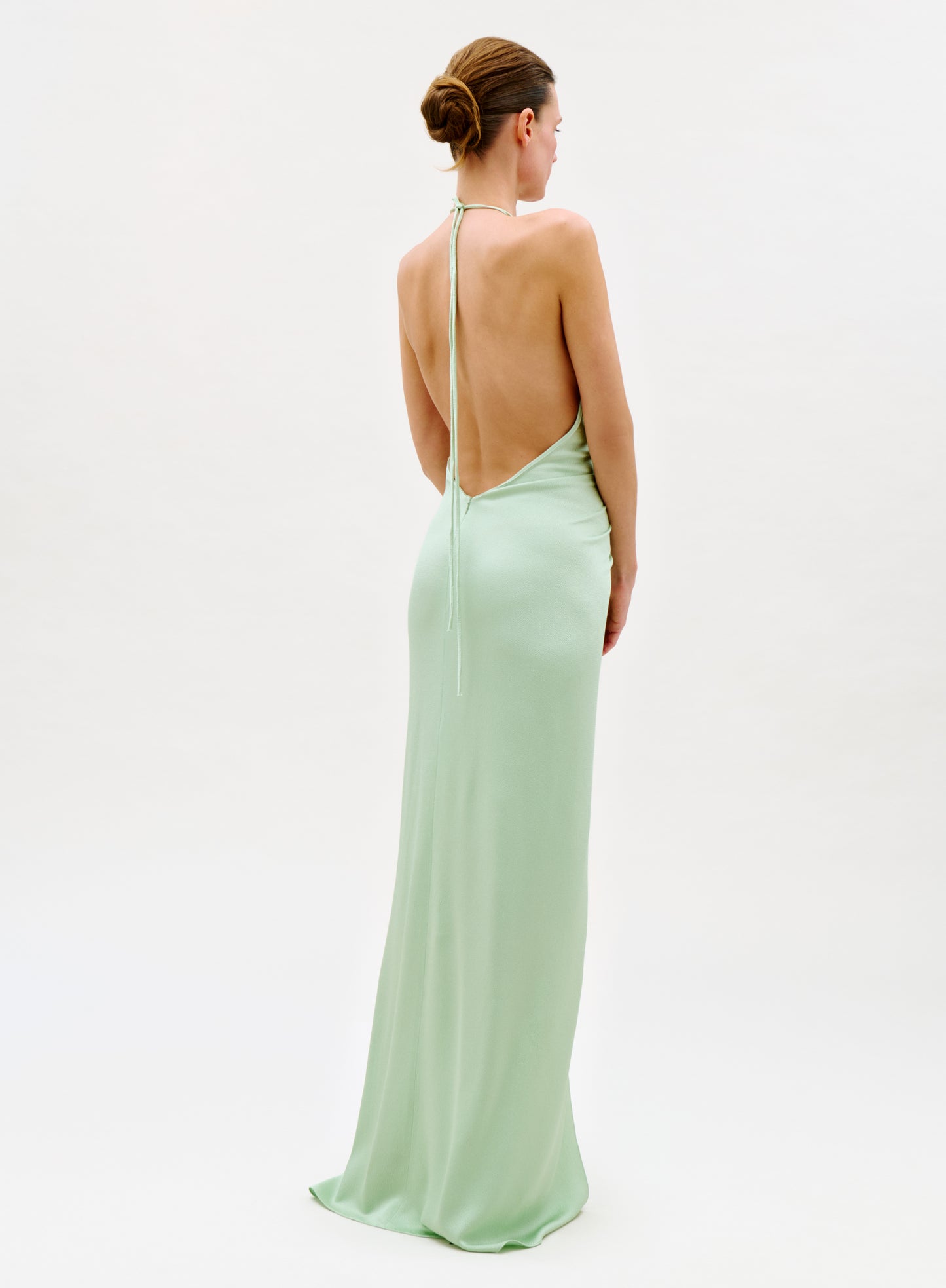 A woman faces away, wearing the Jenifer Dress—a long, light green backless gown with a halter neck tie—her hair in a neat bun against a simple, light background.