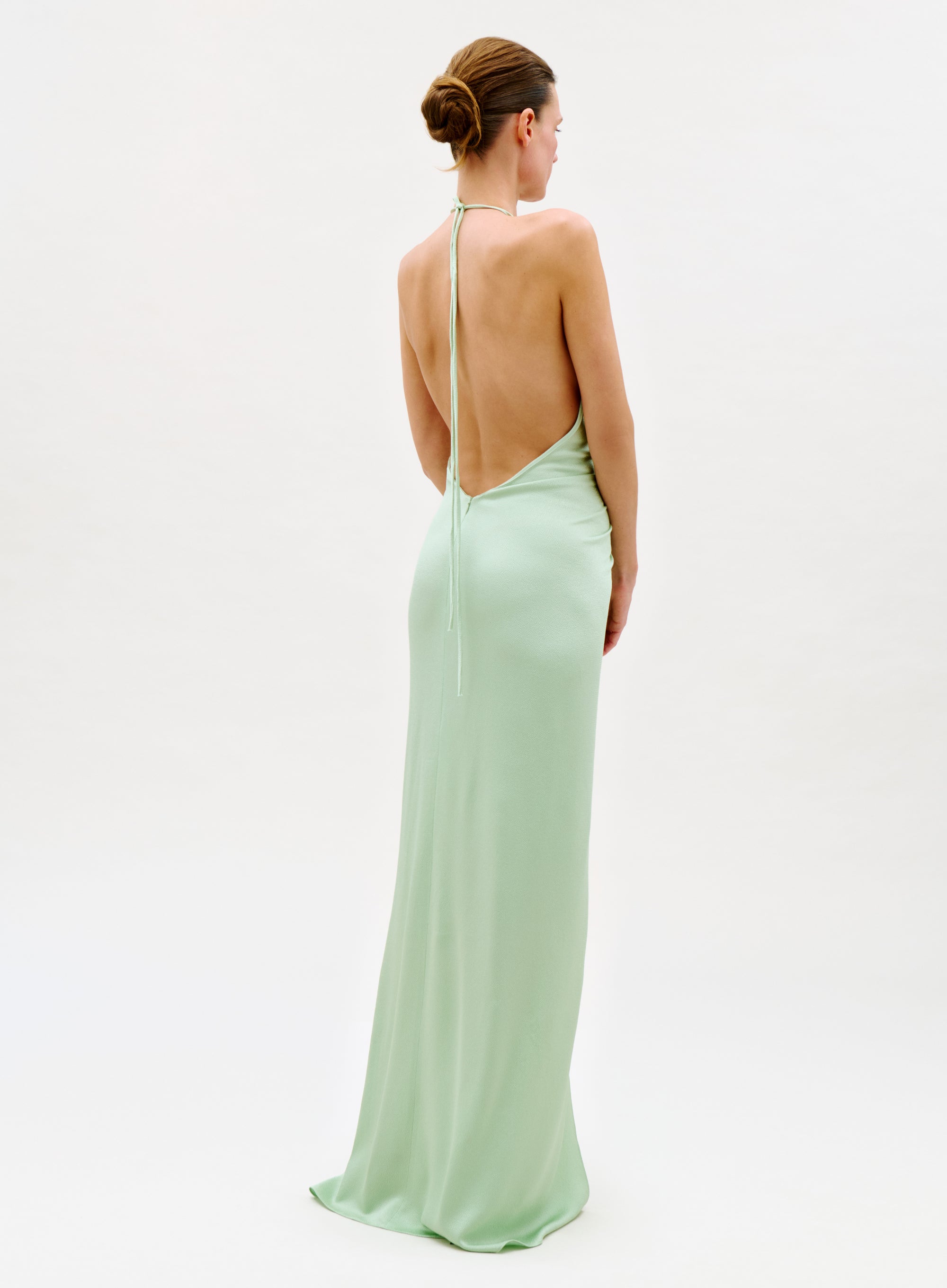 A woman faces away, wearing the Jenifer Dress—a long, light green backless gown with a halter neck tie—her hair in a neat bun against a simple, light background.