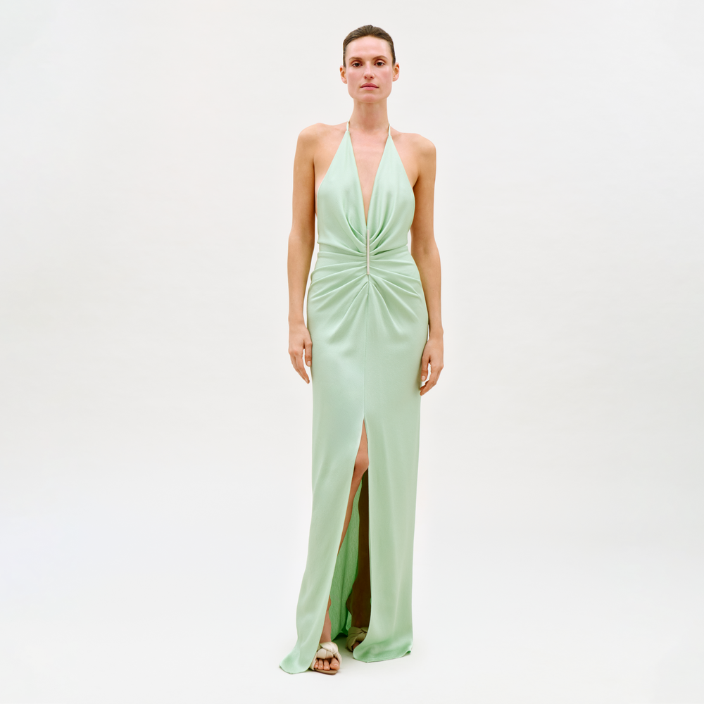 A person stands against a plain white background wearing the Jenifer Dress, a light green sleeveless evening gown featuring a halter neckline, gathered front detail, and a high front slit.
