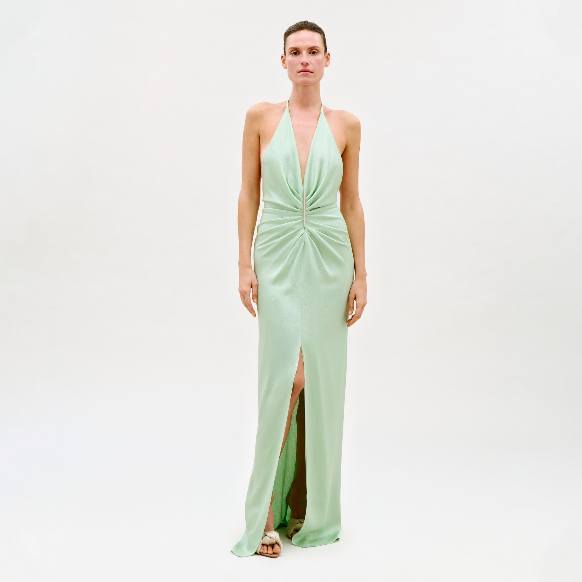 A person stands against a plain white background wearing the Jenifer Dress, a light green sleeveless evening gown featuring a halter neckline, gathered front detail, and a high front slit.