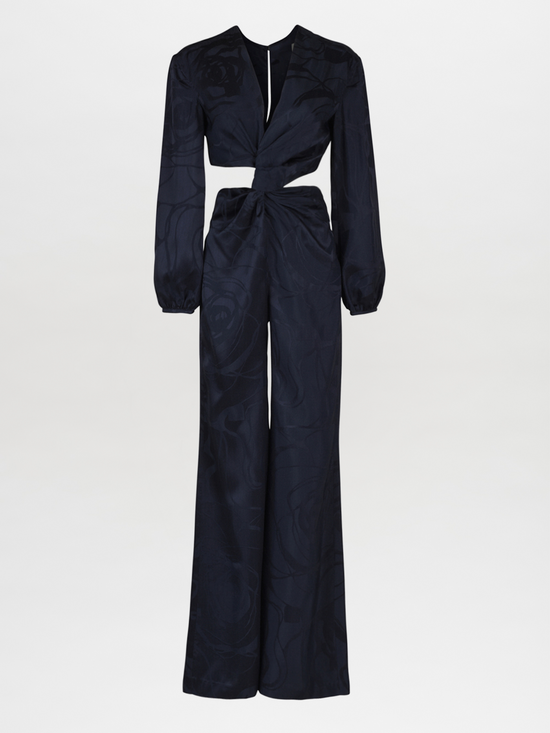 The Joely Jumpsuit Navy Jacquard, a black long-sleeve piece from the Resort 2023 collection, features a twisted front design, cutout at the waist, and wide-leg trousers.