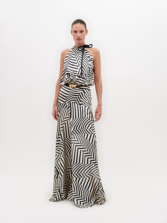 A person is wearing a Joss Dress Beige Black Stripes, accented with a black and gold belt, standing against a plain white background. This ensemble is available for pre-order now and will ship by November 15th, 2024.