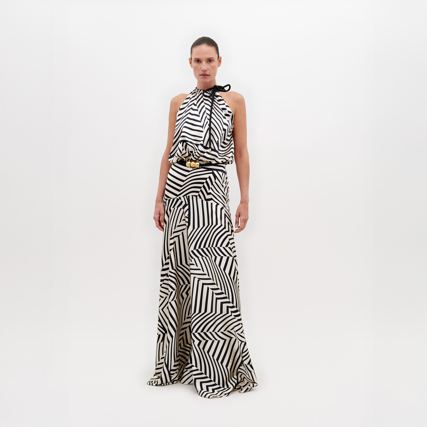 A person is wearing a Joss Dress Beige Black Stripes, accented with a black and gold belt, standing against a plain white background. This ensemble is available for pre-order now and will ship by November 15th, 2024.