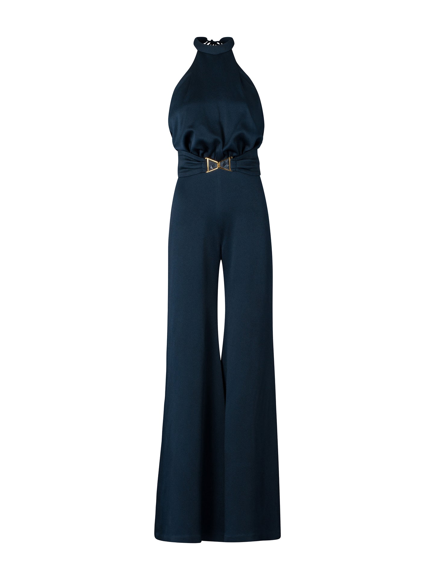 A woman wearing the Kaede Jumpsuit Navy, featuring a halter-neck design with wide-leg pants and a black belt, stands against a light blue background. She accessorizes with multiple black bracelets on one arm, exuding an air of chic nautical elegance.