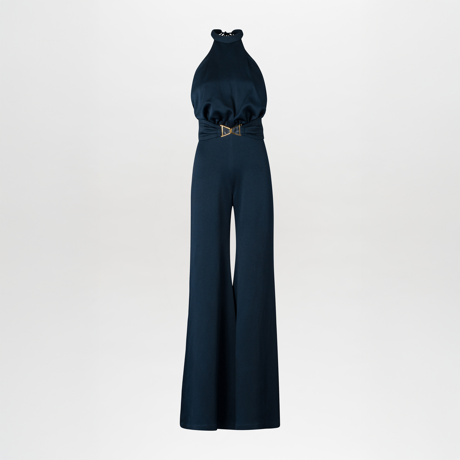 A woman wearing the Kaede Jumpsuit Navy, featuring a halter-neck design with wide-leg pants and a black belt, stands against a light blue background. She accessorizes with multiple black bracelets on one arm, exuding an air of chic nautical elegance.