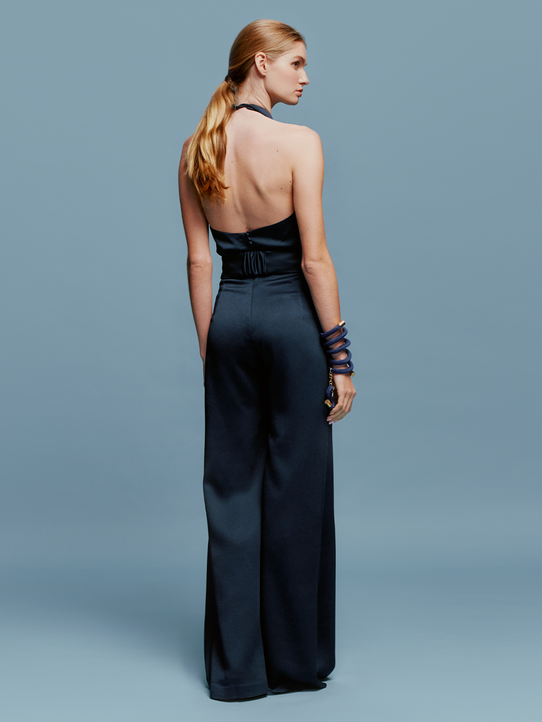 A woman wearing the Kaede Jumpsuit Navy, featuring a halter-neck design with wide-leg pants and a black belt, stands against a light blue background. She accessorizes with multiple black bracelets on one arm, exuding an air of chic nautical elegance.