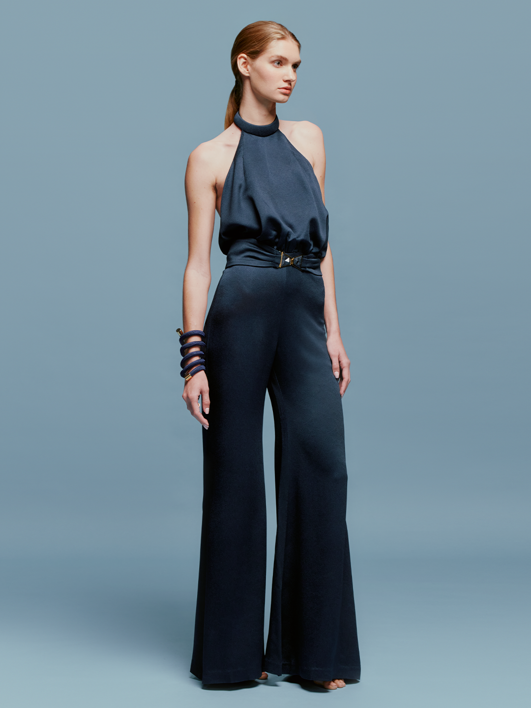 A woman wearing the Kaede Jumpsuit Navy, featuring a halter-neck design with wide-leg pants and a black belt, stands against a light blue background. She accessorizes with multiple black bracelets on one arm, exuding an air of chic nautical elegance.