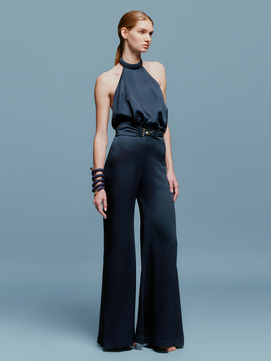 A woman wearing the Kaede Jumpsuit Navy, featuring a halter-neck design with wide-leg pants and a black belt, stands against a light blue background. She accessorizes with multiple black bracelets on one arm, exuding an air of chic nautical elegance.