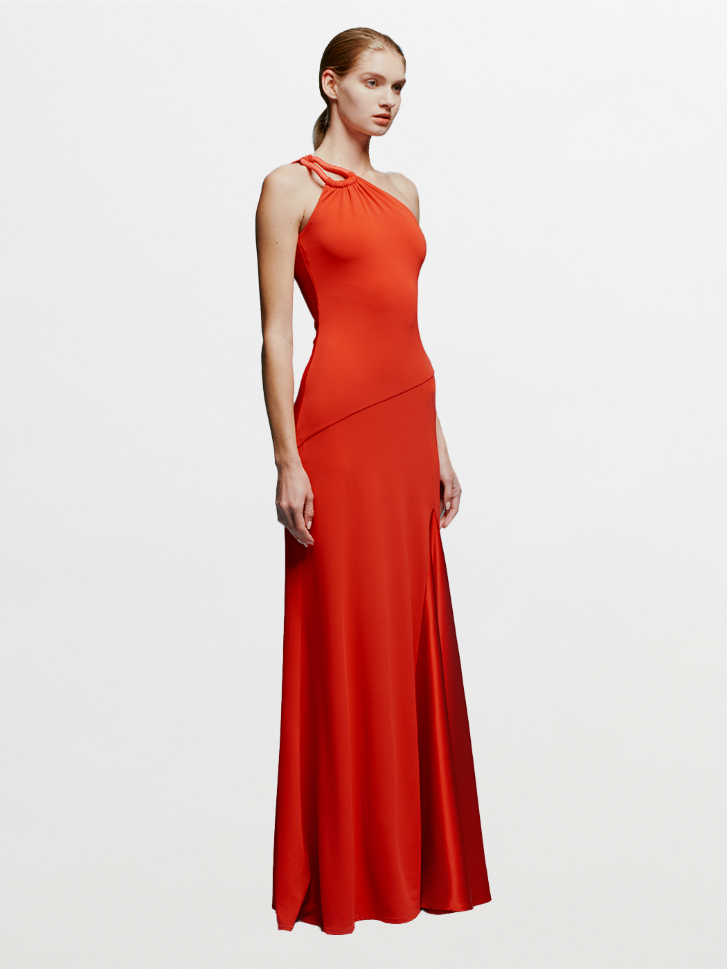 A woman in a Kanya Dress Rouge, featuring a red, one-shoulder design and floor-length cut, stands against a plain blue background. Her hair is pulled back as she calmly stands with her arms by her sides, appearing to wait for an important date such as August 19th or September 2nd.