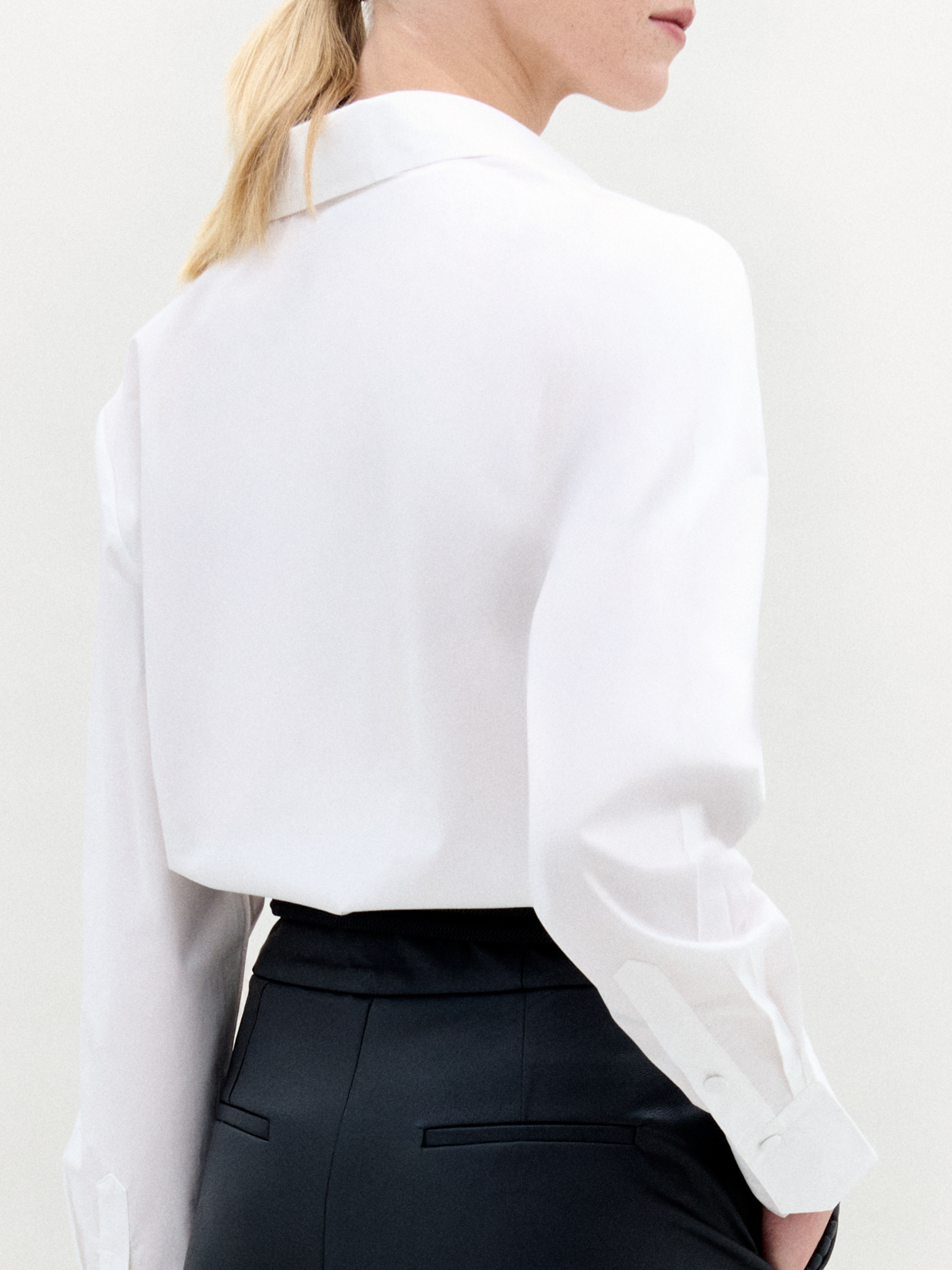 A person with blond hair wears the Karole Blouse, which has long sleeves and floral cutout details, paired with black trousers and a gold belt, standing against a plain white background.