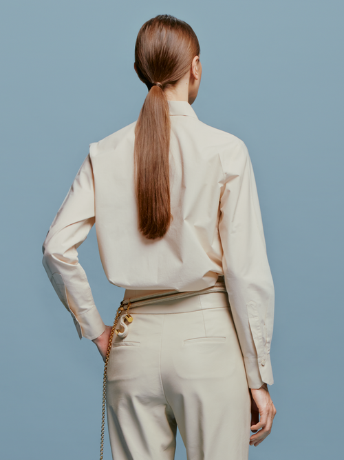 A person stands against a blue background, wearing the Karole Blouse Cream adorned with large floral decorations and holding a small accessory attached to their pants. Please note that this item’s shipping dates are between August 19th and September 2nd.