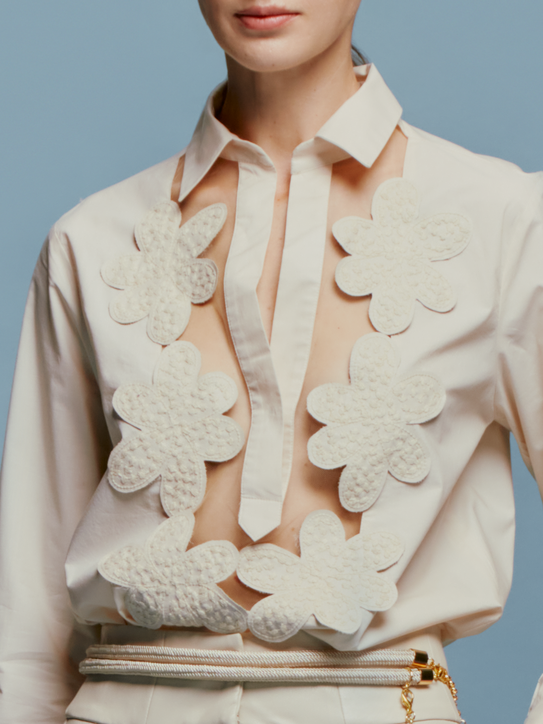 A person stands against a blue background, wearing the Karole Blouse Cream adorned with large floral decorations and holding a small accessory attached to their pants. Please note that this item’s shipping dates are between August 19th and September 2nd.